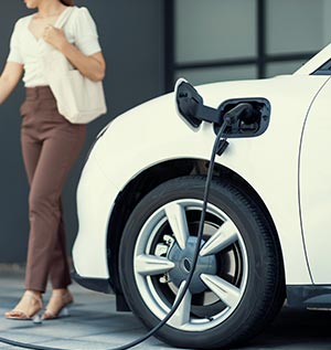 Woman walking near a white electric vehicle charging at a station, showcasing Carter Services' electric vehicle charging installation expertise.