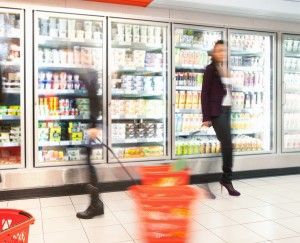 Woman shopping in a grocery store aisle with commercial refrigeration units displaying various products in the background, emphasizing the importance of reliable refrigeration for food safety.